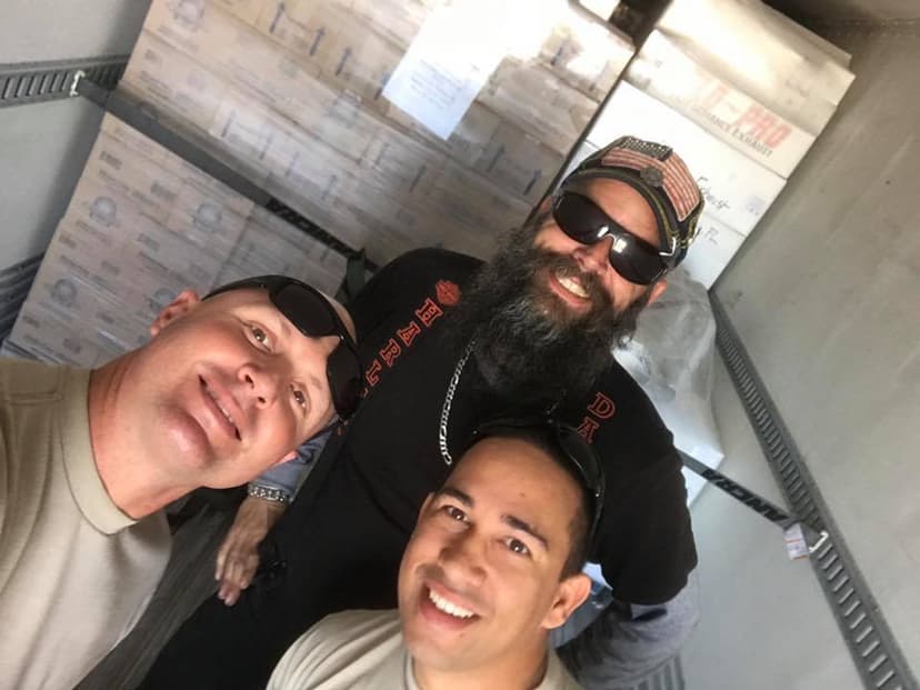 Three crew members selfie inside the trailer, walls of Kinnikinnick boxes stacked behind them