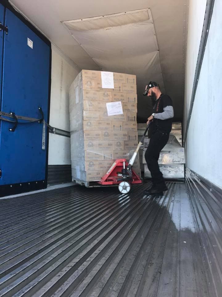Crew member wearing a headlamp pushing a tall pallet of Kinnikinnick boxes with a red pallet jack inside a trailer