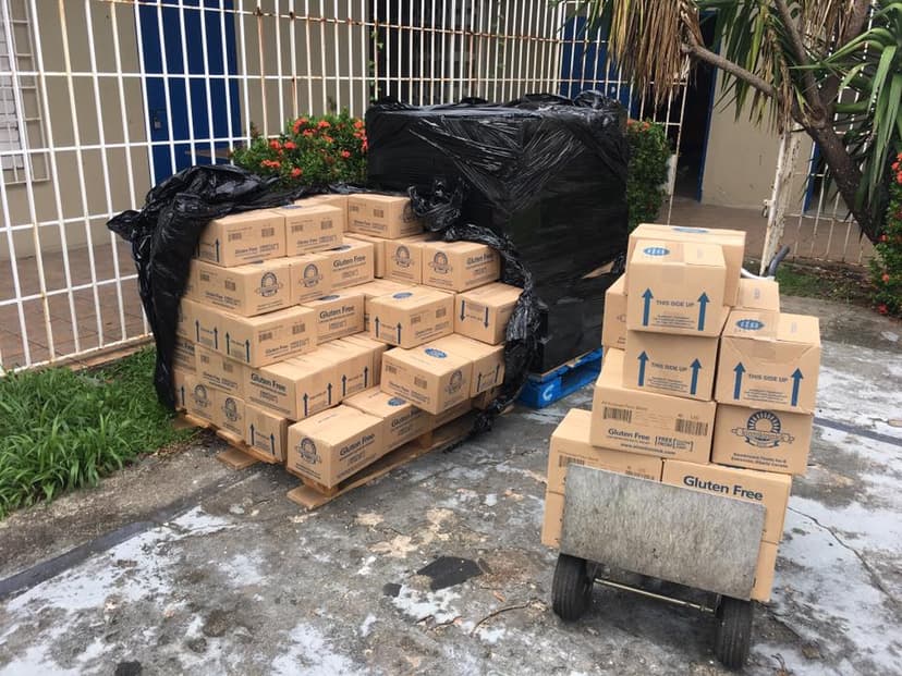 Kinnikinnick Gluten Free boxes unloaded on pallets and a hand cart outside the Alianza de Autismo building, iron fence and flowering plants behind
